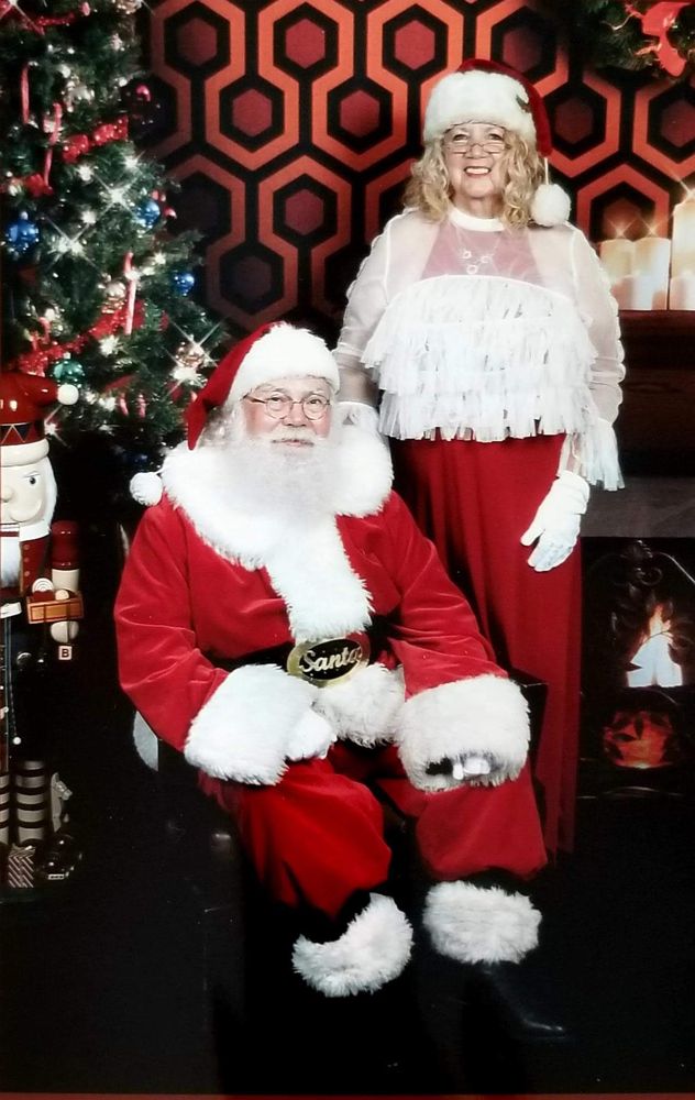 Santa John seated with Mrs. Claus standing beside him in front of a Christmas tree